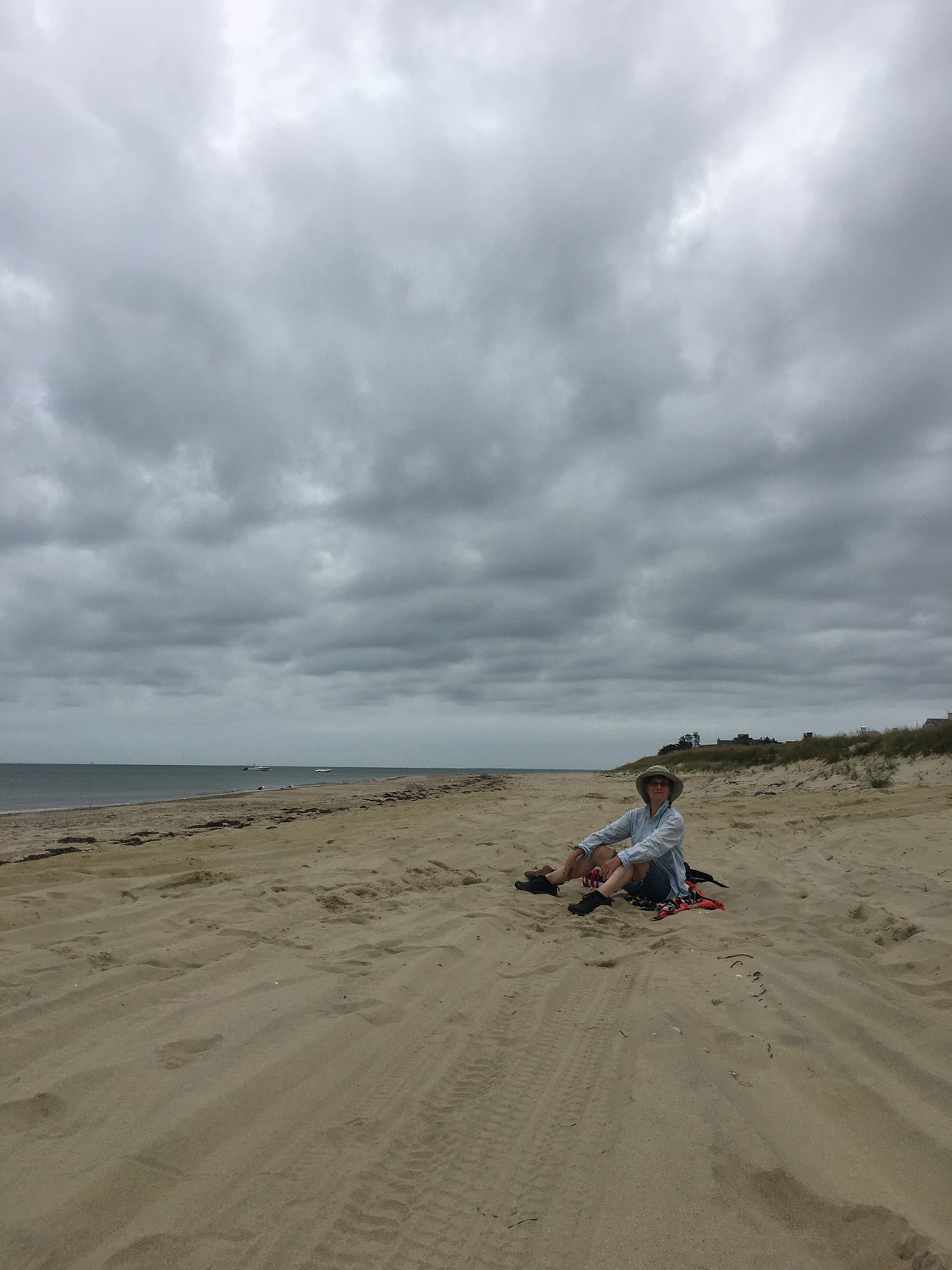 The artist, small in the frame, sitting on a wide Nantucket beach under an enormous grey weather-sky, the sea to the left