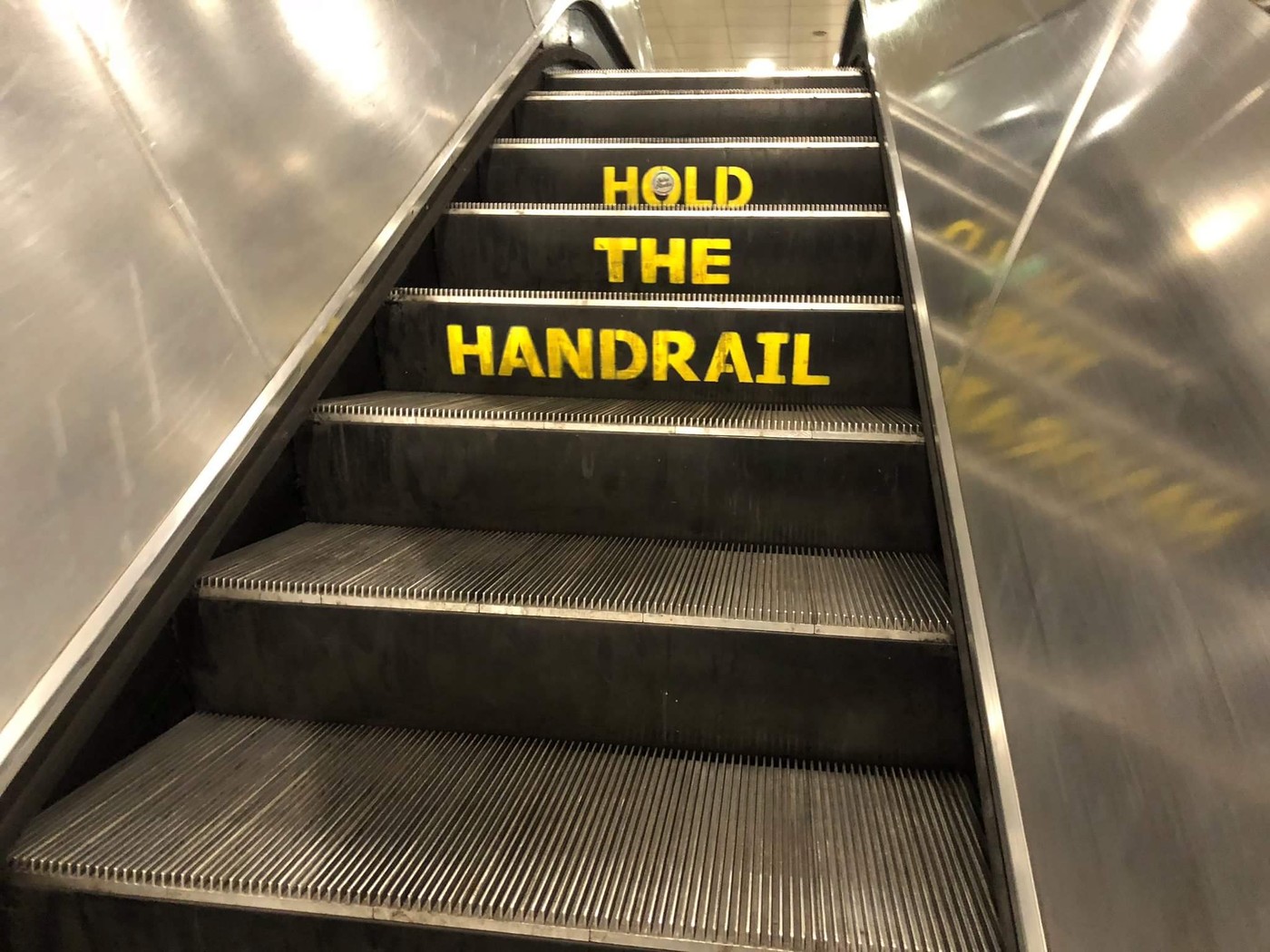 Looking up the steps of a metal subway escalator; three steps read, in yellow stencil, HOLD THE HANDRAIL